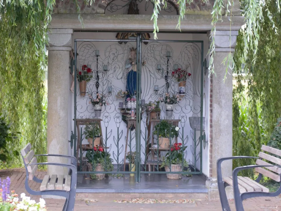 Kapel met beeld van Maria achter sierlijk hekwerk, omringd door bloemen en hangende wilgentakken in Vlodrop, onderdeel van de gelukswandeling in Vlodrop