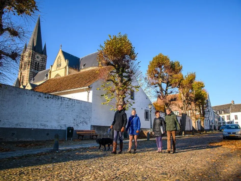 Een groep mensen wandelt met een hond over een geplaveide straat in een historisch dorp, met een kerk en witgekalkte gebouwen op de achtergrond op een zonnige dag.