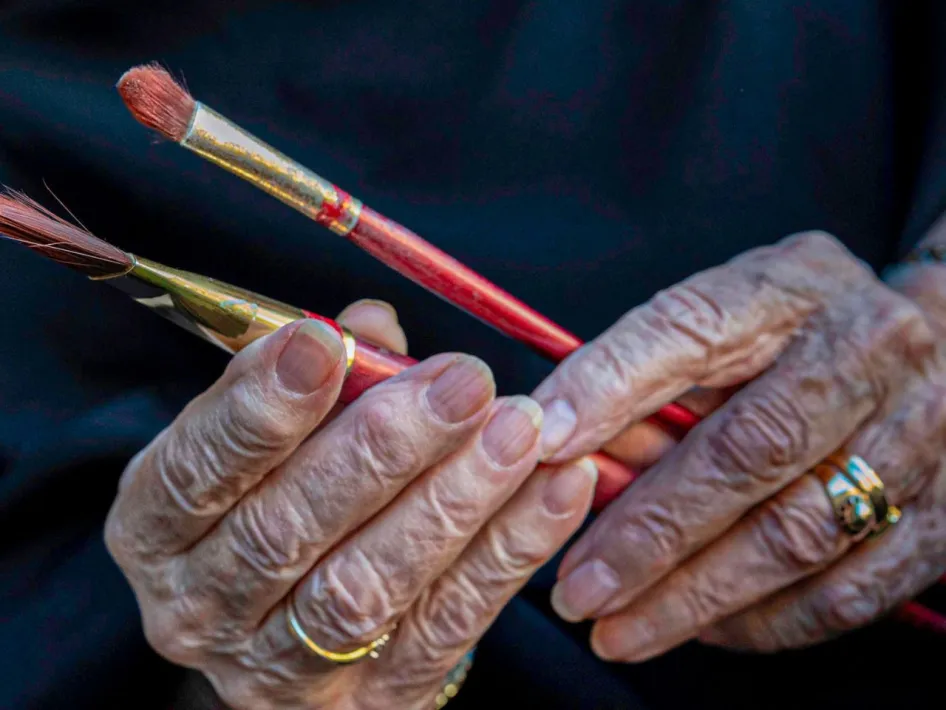 Twee oudere handen houden rode penselen vast tegen een donkere achtergrond.