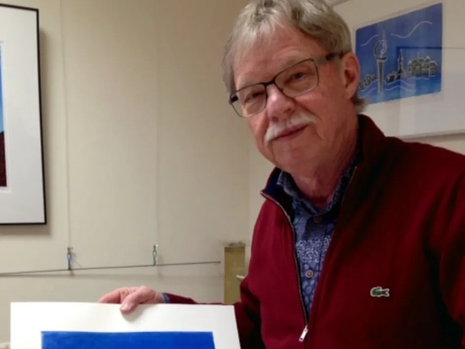 Man met bril en rode trui toont een blauwe grafische afdruk in een galerie- of atelieromgeving.