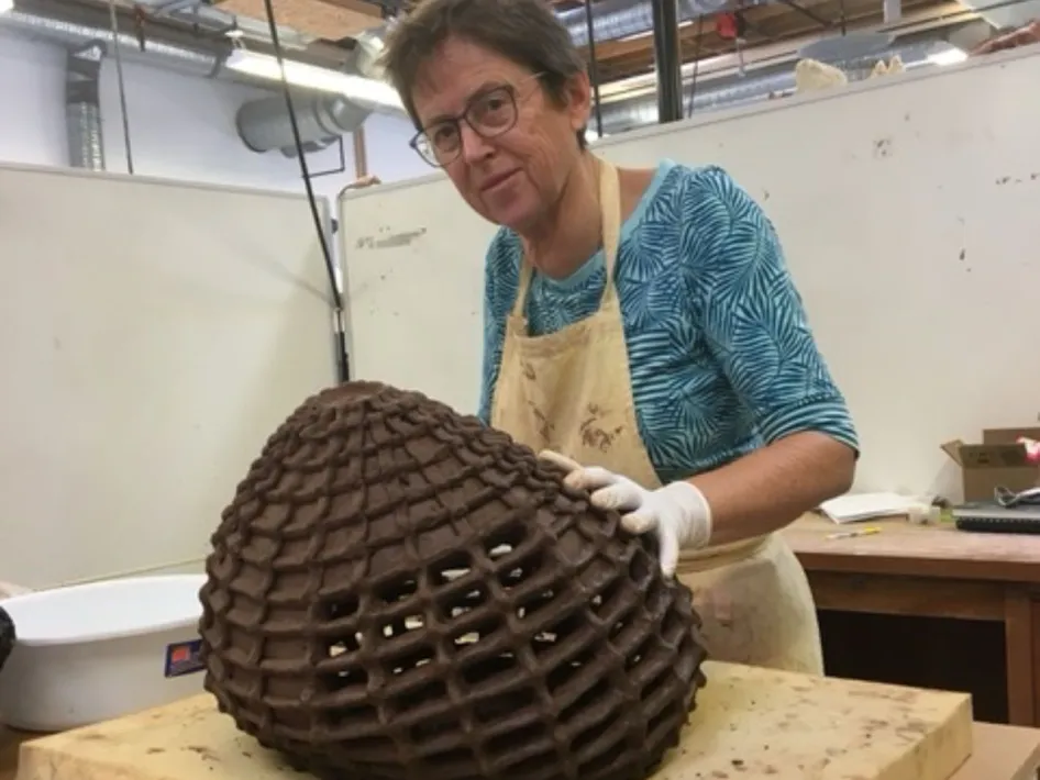 Vrouw met bril en schort werkt aan een groot, opengewerkt keramisch object in een atelier.