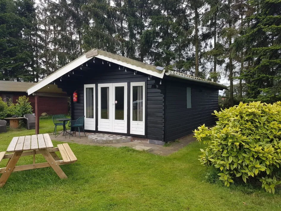 Zwarte houten blokhut met veranda, witte deuren en tuinmeubilair, gelegen in een groene, bosrijke omgeving met picknicktafel op het grasveld.