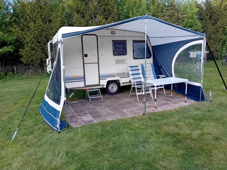 Kleiner Wohnwagen mit Vorzelt und Gartengarnitur auf einer gefliesten Terrasse, gelegen auf einer grünen Campingwiese mit Bäumen im Hintergrund.