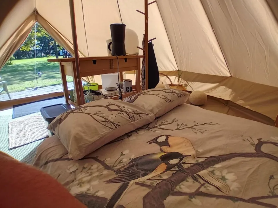 Interior of a tastefully decorated tent overlooking a lawn, with a made-up bed with a bird motif and a wooden table with camping equipment.