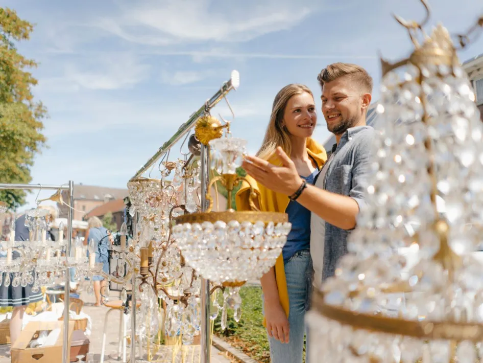 Stel bekijkt kristallen kroonluchters op een zonnige vrijmarkt, met andere bezoekers en kramen op de achtergrond.