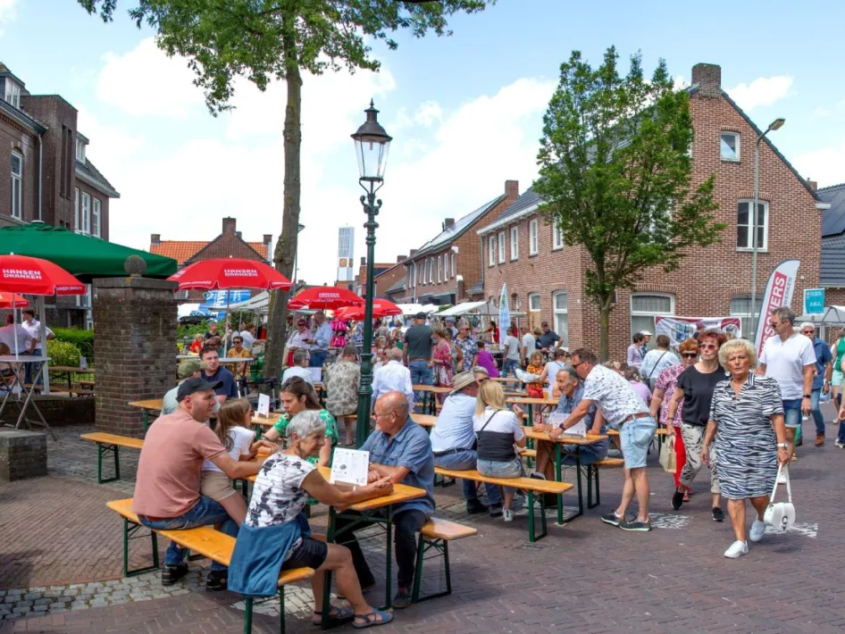Drukke zomerse braderie in een dorp, met mensen aan picknicktafels en kraampjes langs de straat onder rode parasols.