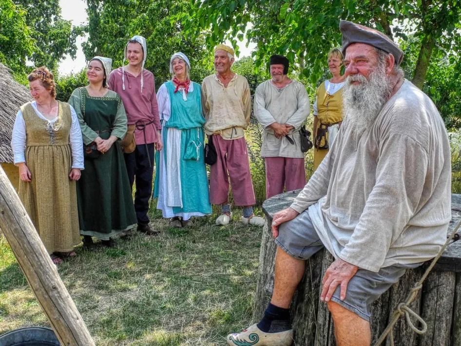 Groep mensen in middeleeuwse kleding tijdens een historisch evenement, met op de voorgrond een man met grijze baard zittend op een boomstam, en op de achtergrond lachende deelnemers in kleurrijke kostuums.