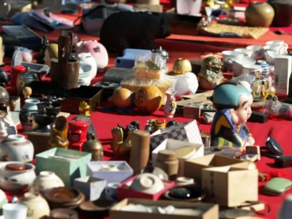 Uitzicht op een rommelmarkttafel vol tweedehands spulletjes, curiosa, serviesgoed en kleine beeldjes, uitgestald op een rood tafelkleed.