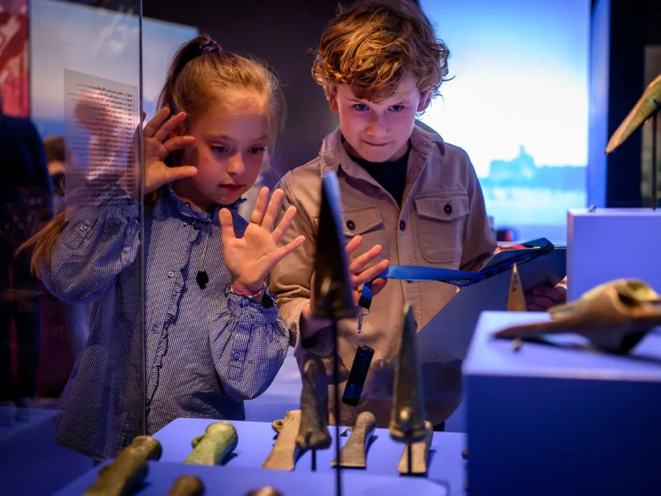 Twee kinderen kijken gefascineerd naar bronzen voorwerpen in een vitrine tijdens de tentoonstelling Betoverend Brons.
