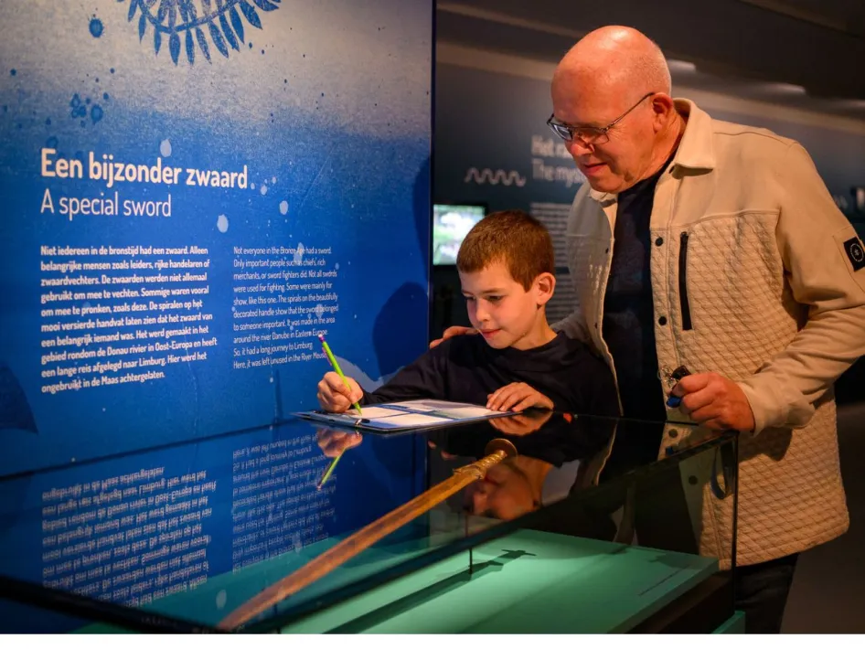 Man en kind bekijken samen een vitrinekast met een bijzonder zwaard, terwijl ze aantekeningen maken op een invulblad.