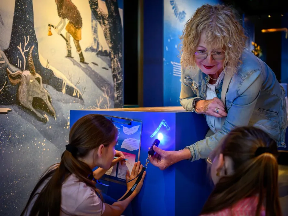 Een vrouw laat twee kinderen iets ontdekken met een blauw lampje bij een interactieve opstelling in de tentoonstelling.