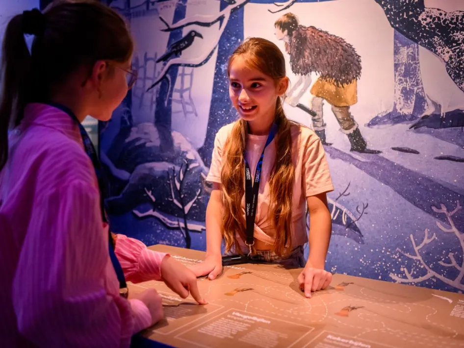 Twee meisjes bekijken samen een interactieve kaart in de tentoonstelling, met een getekende wand vol winterse bosfiguren op de achtergrond.