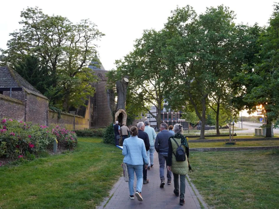 Groep mensen tijdens een avondwandeling langs historische stadsmuur en oude kerktoren, omringd door bomen en bloemen, met ondergaande zon op de achtergrond.