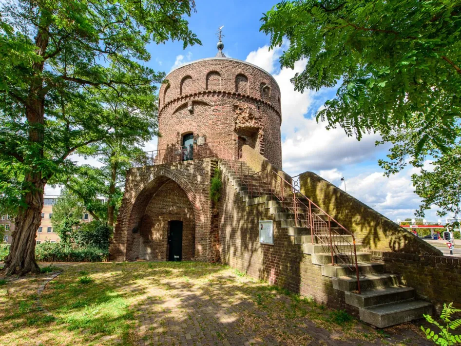 Der Rattenturm in Roermond, ein mittelalterlicher runder Backsteinturm mit Treppe und Torbogen, umgeben von Bäumen und gelegen in einer parkähnlichen Umgebung.