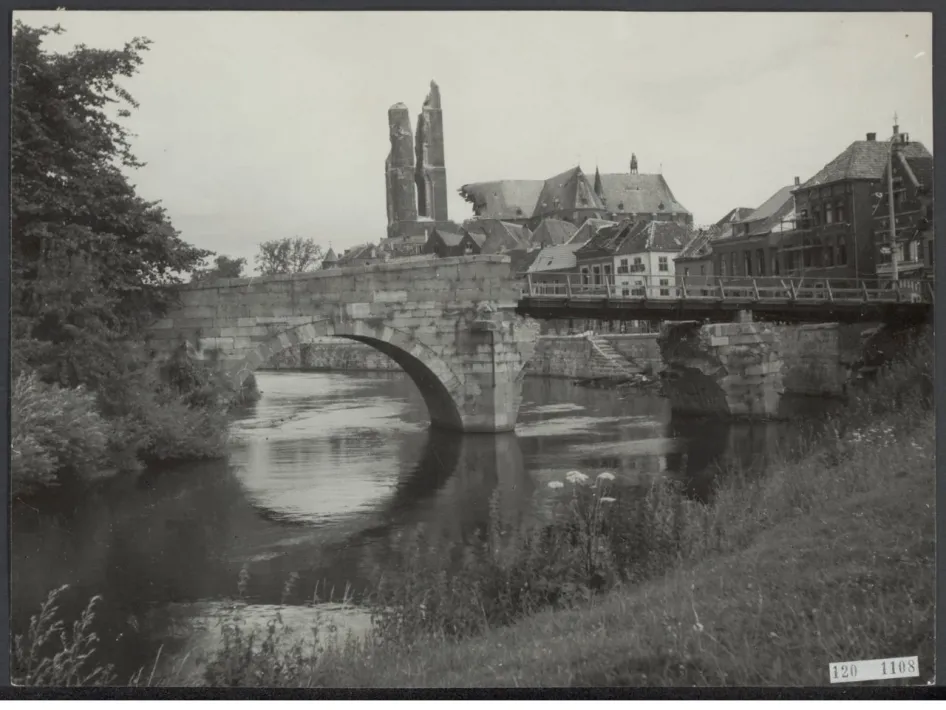 Zwart-witfoto van een historische stenen boogbrug over een rivier, met op de achtergrond een beschadigde kerk met twee torens en oude gebouwen in een stad. Rechts op de brug is een tijdelijke houten constructie zichtbaar.