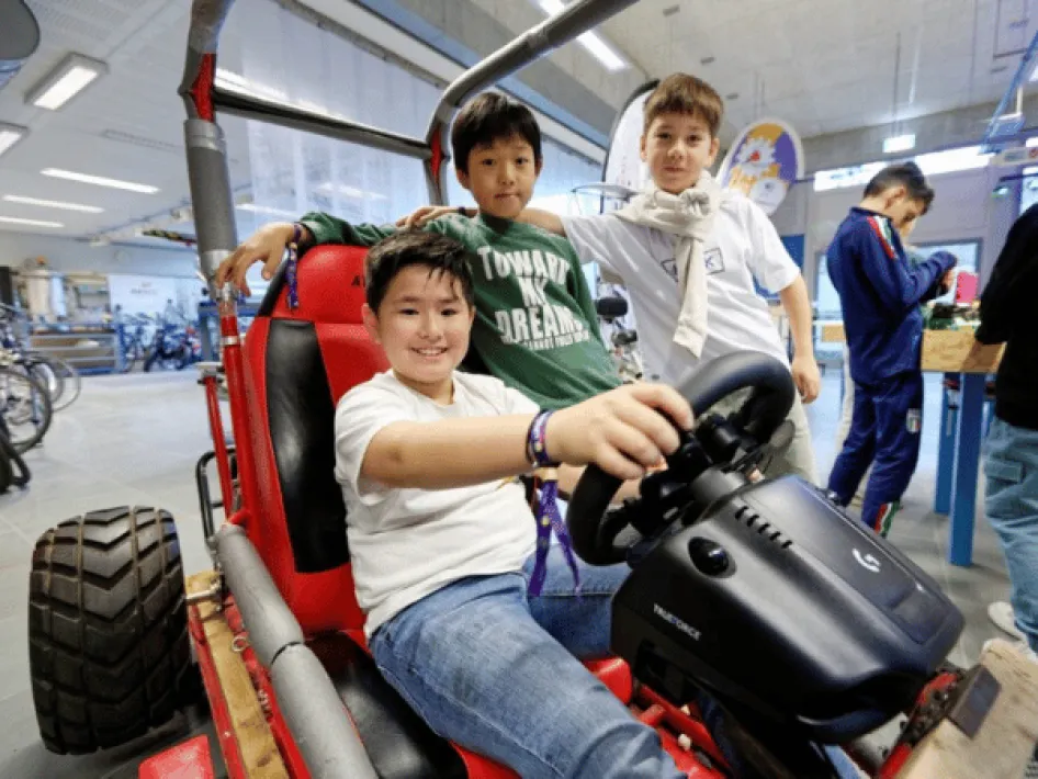 One boy is sitting in the race car, with two other boys standing next to it.