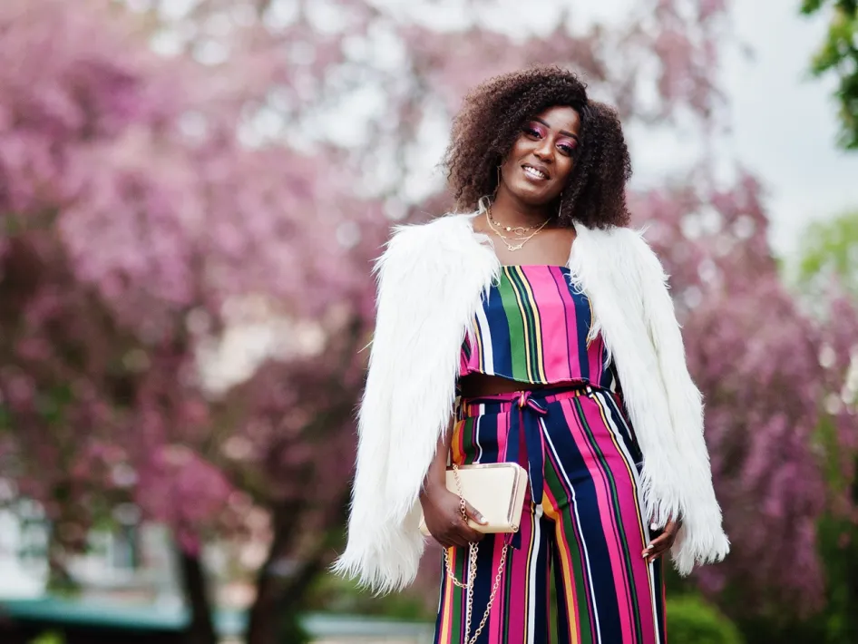 Een vrouw in een kleurrijke gestreepte outfit met een witte fluffy jas poseert glimlachend voor bloeiende roze bomen. Haar stijlvolle look past perfect bij de lente- en zomermode die te zien is tijdens het Fashion Event in Roermond.