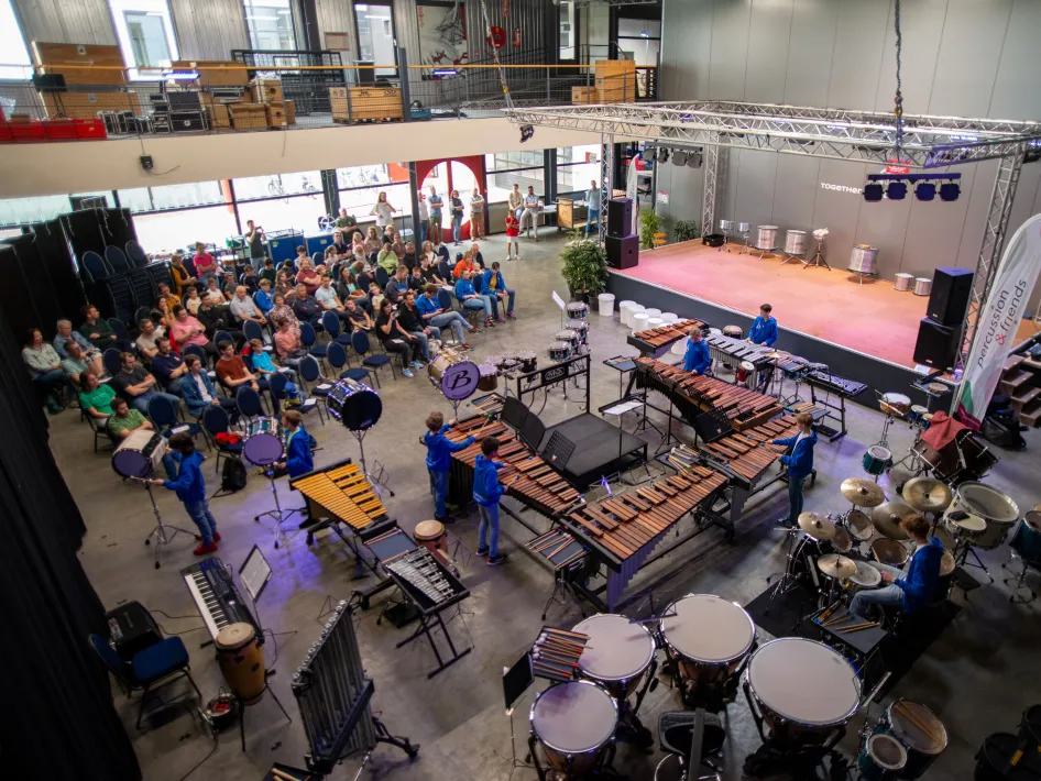 Een muziekvoorstelling in een zaal, met een groot publiek dat kijkt naar een groep muzikanten die op marimba’s, drums en slagwerkinstrumenten speelt.