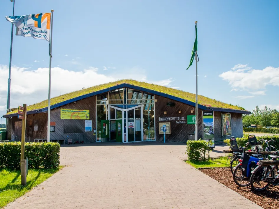 Buitencentrum De Pelen mit Holzfassade, grünem Eingangsbereich, Fahnen und abgestellten Fahrrädern am Eingang des Nationalparks De Groote Peel
