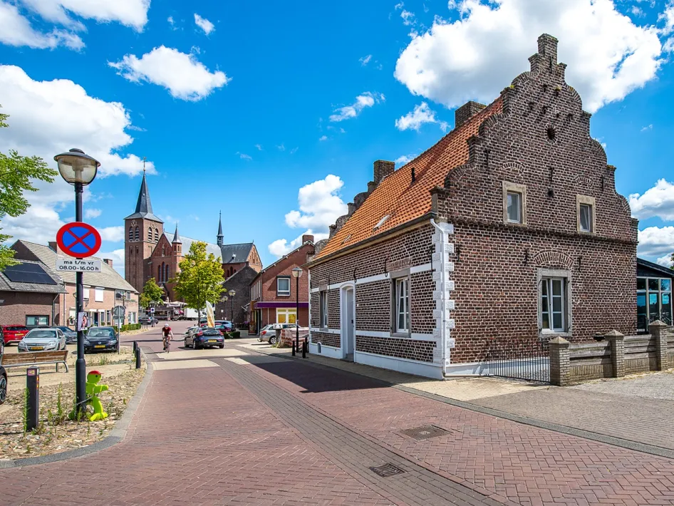 Een dorpsstraat in Neer met oude bakstenen huizen, een rode pannendak en op de achtergrond een grote kerk met meerdere torens onder een blauwe lucht met wolken.