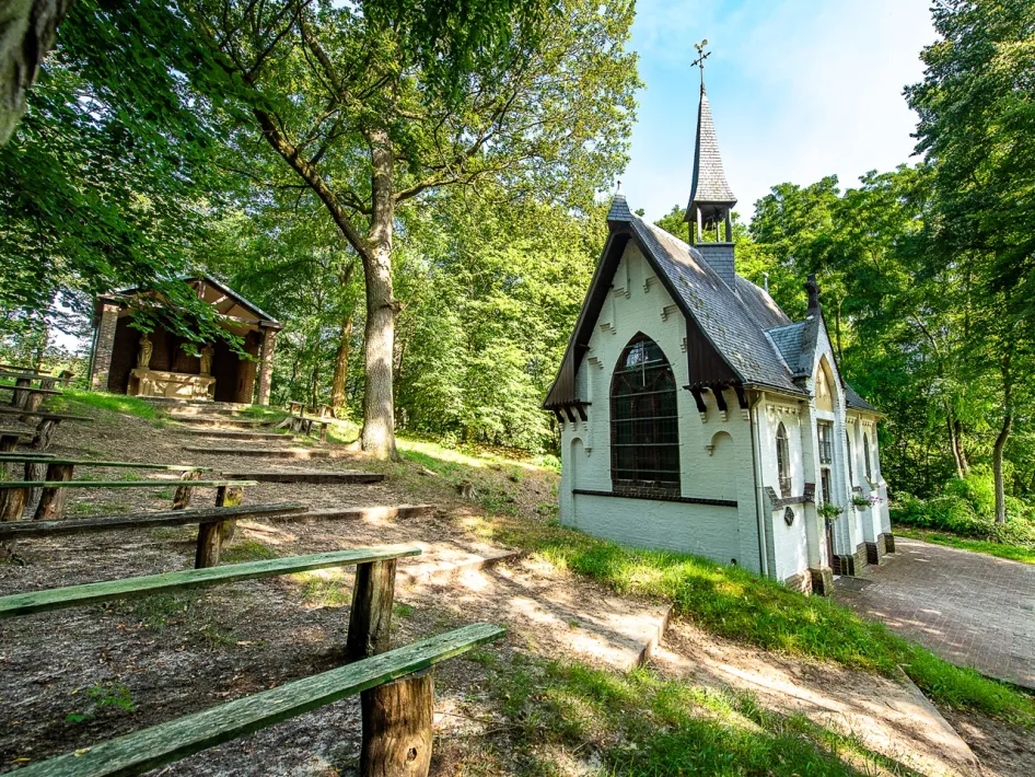 Een witte kapel met hoge ramen en een spits dak, gelegen in een bosrijke omgeving met houten zitbanken op een helling ervoor.