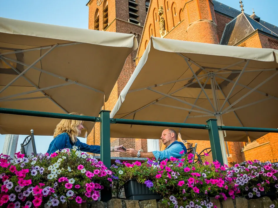 Two people sit on a flower-filled terrace along the Leudal Cycling Route, with a large brick church and beige parasols in the background.