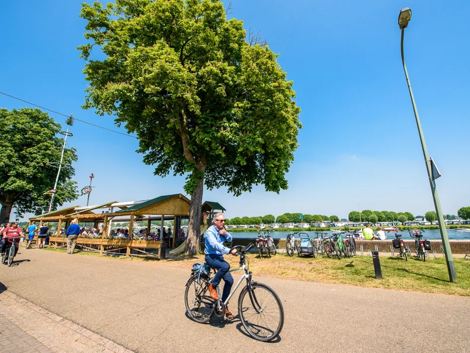 Een zonnige dag bij Café Kanters, waar fietsers en wandelaars genieten van een tussenstop aan het water. Een man fietst voorbij, terwijl andere bezoekers ontspannen op het terras.