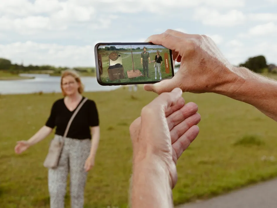 Een persoon houdt een smartphone vast met de Archeo Route Limburg-app, waarop Archie de Archeoloog en een historische scène in augmented reality te zien zijn. Op de achtergrond poseert een vrouw met een rivierlandschap tijdens de Ode aan de Maas fietsroute.
