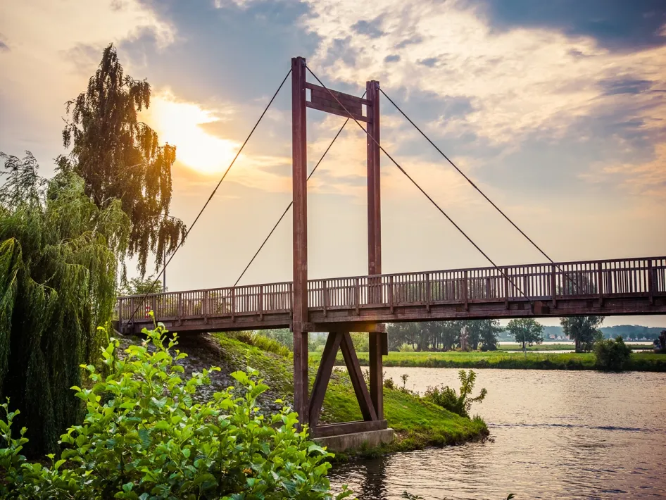 Houten fietsbrug over de Maas bij zonsondergang.