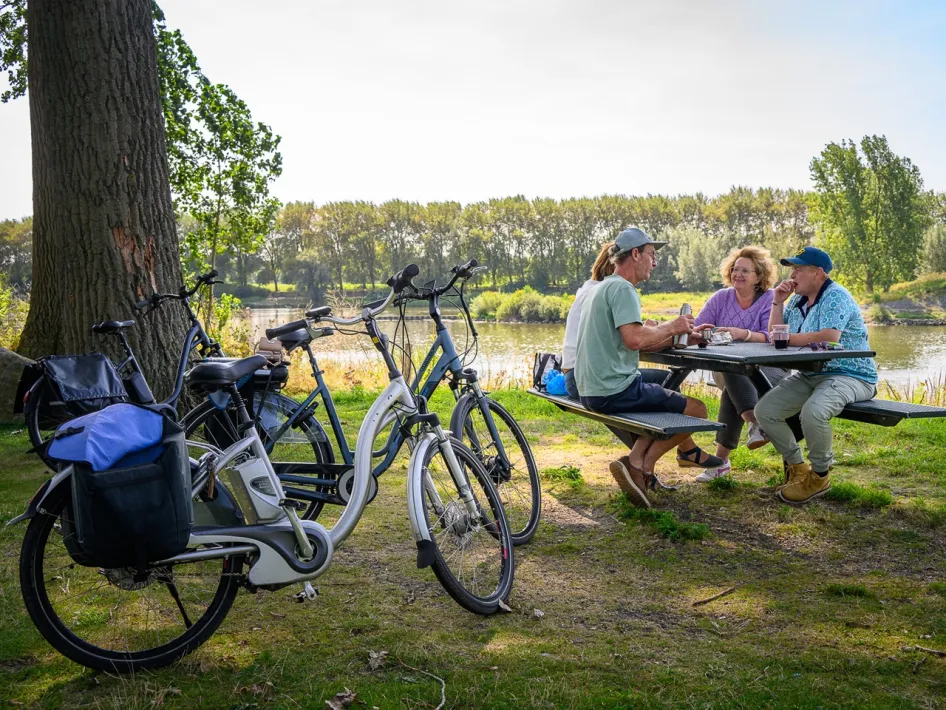 Fietsers pauzeren bij een picknicktafel aan het water, met hun fietsen onder een boom.