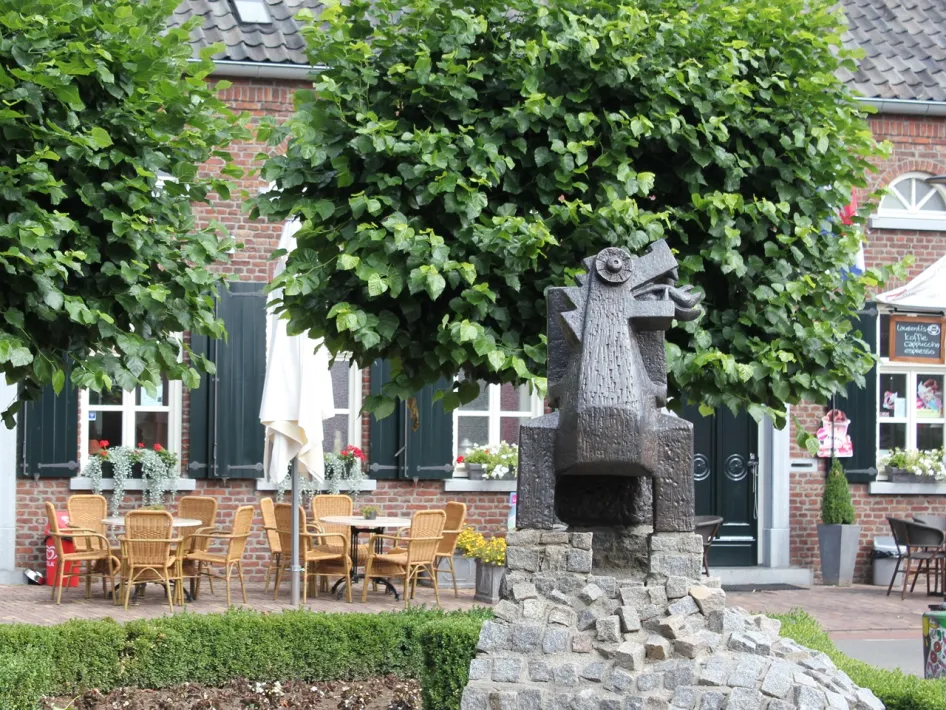 Donker standbeeld van een draak op een stenen sokkel voor een terras met stoelen en bakstenen gevels met groene luiken.