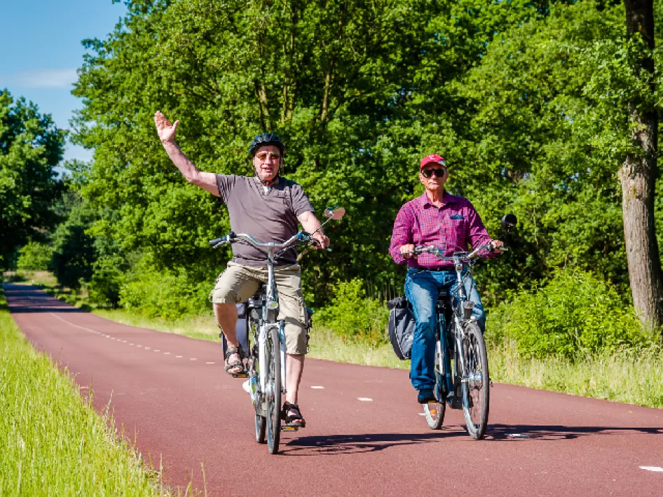 Twee deelnemers aan de Fietsvierdaagse de Roerstreek fietsen over een rustig, rood fietspad, omringd door groene bomen. Eén van hen zwaait enthousiast naar de camera.