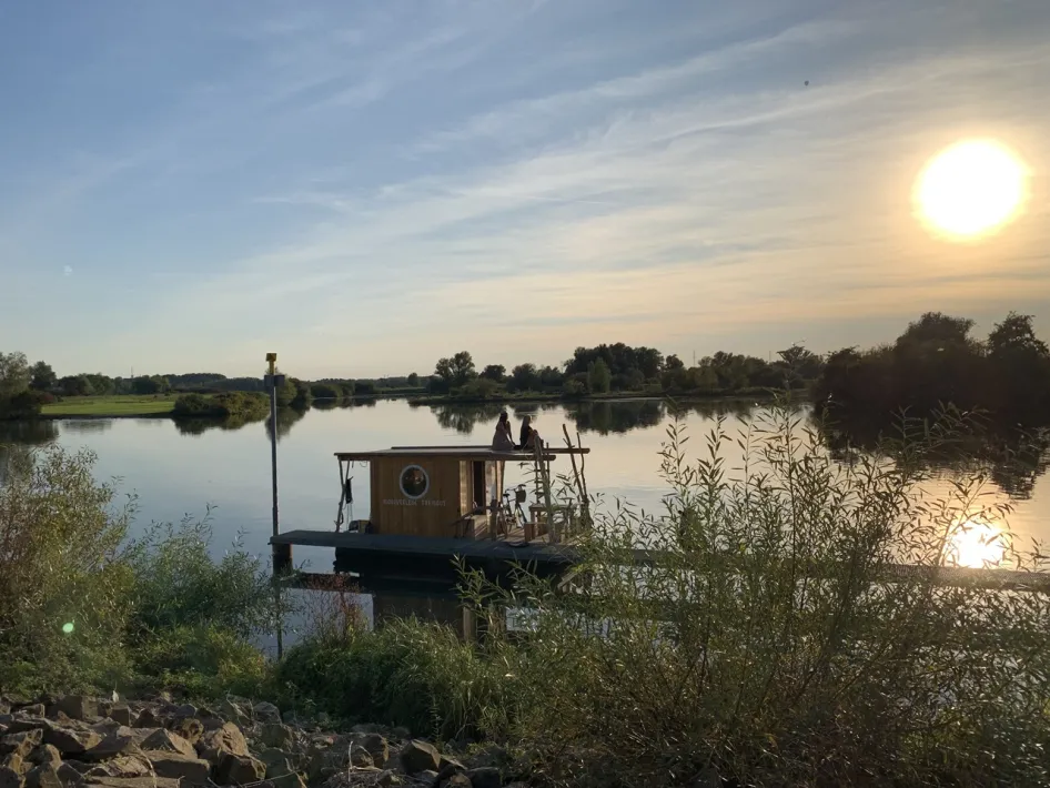 Two people sit on a floating raft from Flossverleih TreibGuT on the Maas, overlooking the water and surrounding landscape.