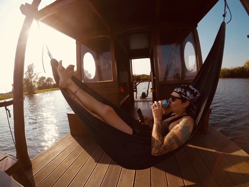 Eine Person liegt in einer Hängematte auf einem Floß auf dem Wasser, mit Sonnenlicht und Blick über die Maas.