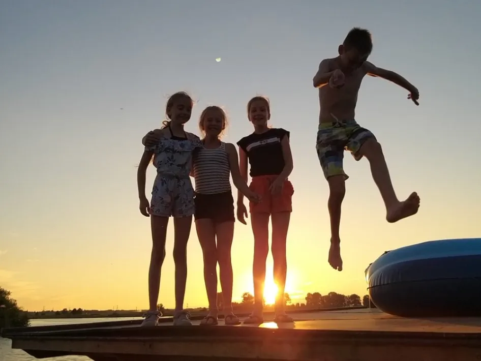 Kinderen springen van een kampeervlot het water in bij zonsondergang.