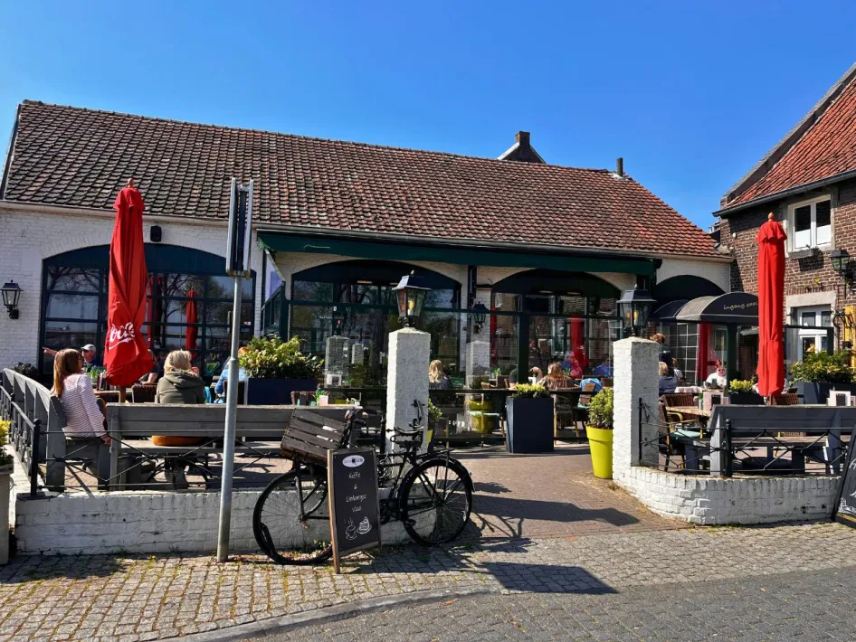 Cosy terrace at Gasterie De Knip with red parasols and guests enjoying the sun beside a white building with a red roof.