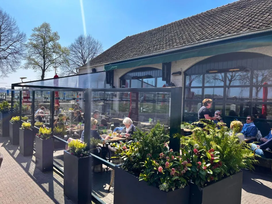 Zonnig terras met glazen windschermen en bloembakken bij Gasterij de Knip, met uitzicht op de Maas en genietende gasten.