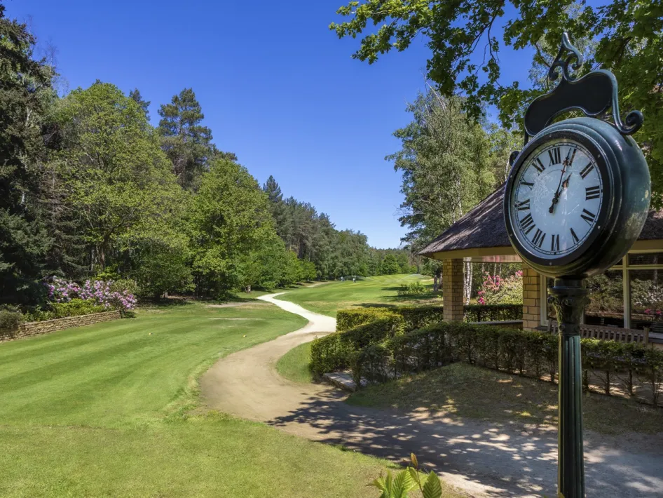 Sierlijke klok bij het clubhuis van golfbaan De Herkenbosche, met uitzicht op een bosrijke fairway.