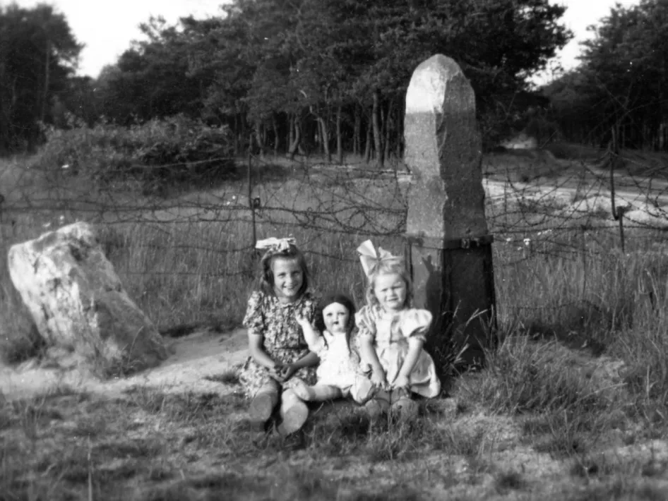 Zwei Mädchen und eine Puppe posieren an einem Grenzpfosten mit Stacheldraht bei De Witte Stein, einem Ort, an dem Grenze und Kriegsgeschichte spürbar aufeinandertreffen.