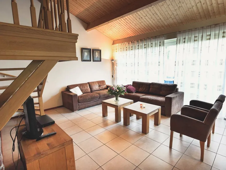 Woonkamer met leren banken, houten tafels en een trap naar boven onder een houten plafond.