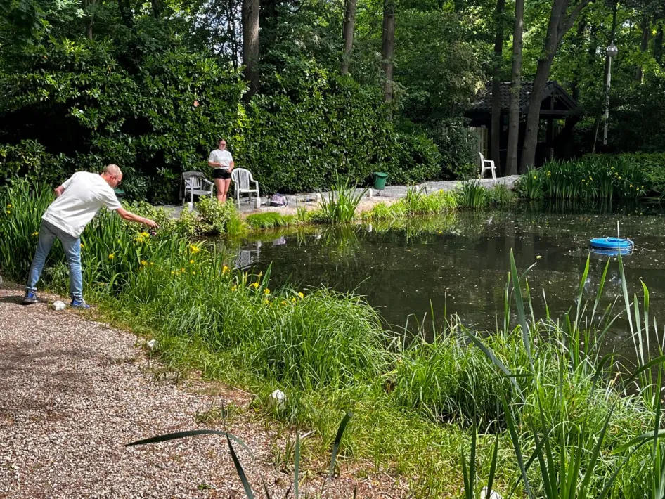 A man and a woman fishing peacefully by a green pond, surrounded by forest and blooming yellow flowers.
