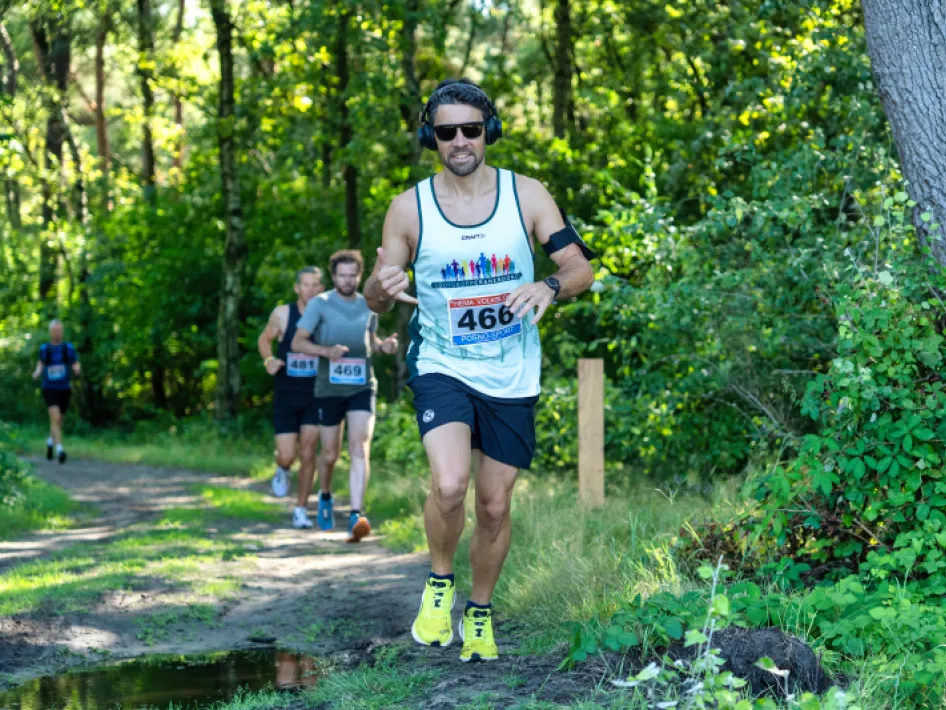 Hardlopers tijdens een trailrun door een bosrijk gebied, met een deelnemer op de voorgrond (rugnummer 466) die een witte singlet draagt en oordopjes en een zonnebril op heeft.