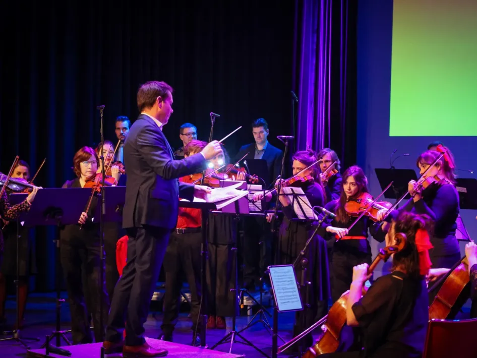 Dirigent en orkest van Heritage Sinfonietta tijdens een concert op een podium met paarse en groene belichting.