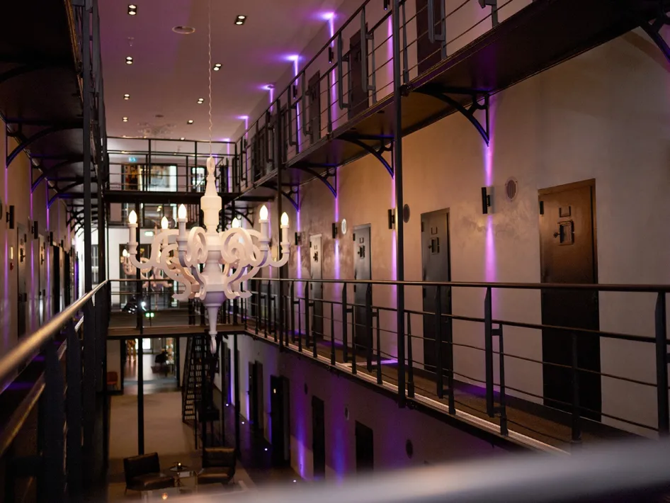 Voormalige cellengang van Het Arresthuis in Roermond, met galerijen, celdeuren, een grote witte kroonluchter en paarse sfeerverlichting.