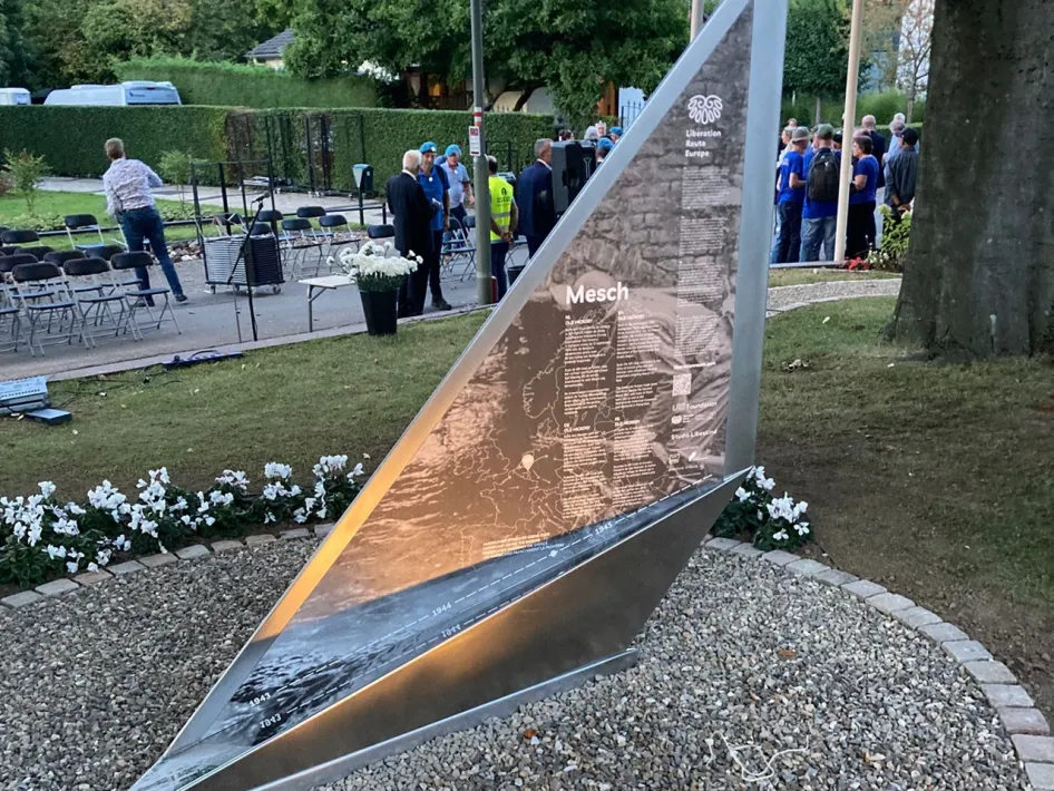 A modern, triangular plaque with information on the liberation of Mesch, part of the Hiking Trail Limburg.