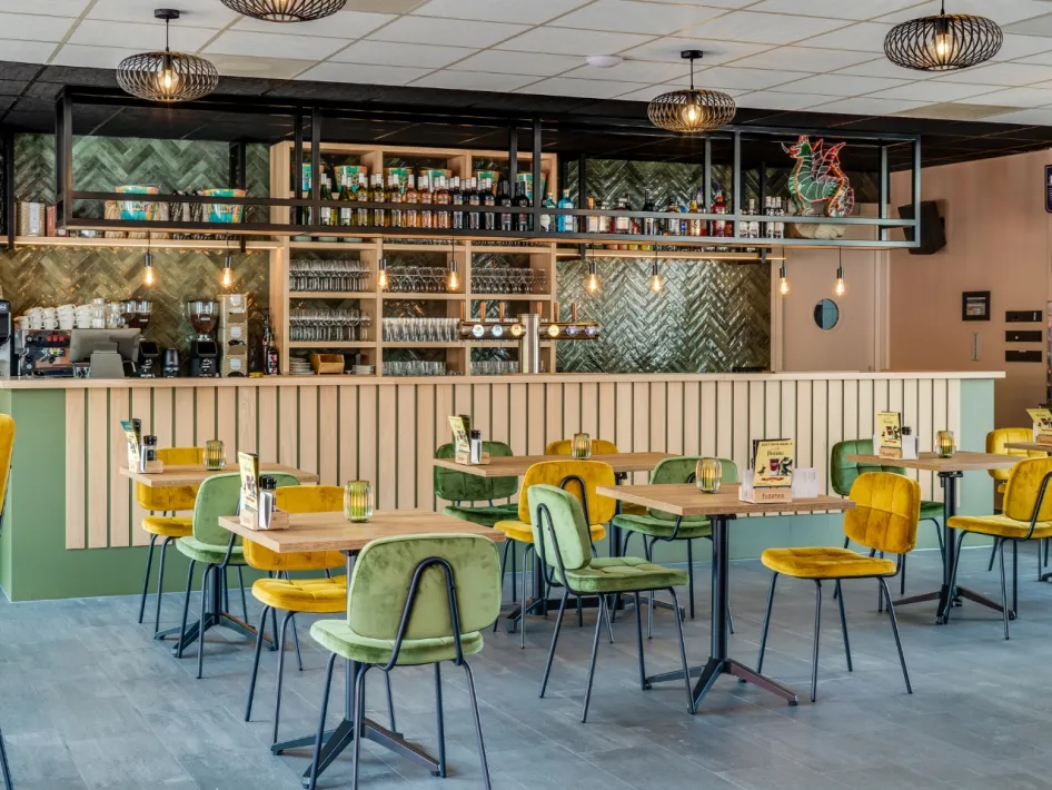 Sfeervol ingerichte ruimte met groene en gele stoelen, houten tafels en een moderne bar met glazen en flessen op de achtergrond.