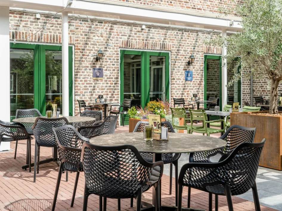 Sunny terrace with black chairs and round tables, located on a brick facade with green doors.