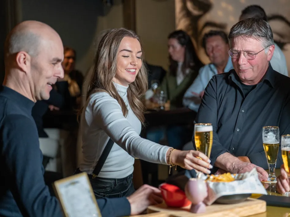 A waitress serves beer and snacks to two men in a cozy bar, while other guests enjoy their evening in the background.
