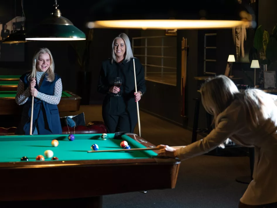 Drie vrouwen spelen pool in een sfeervolle setting. Twee van hen kijken lachend toe terwijl de derde zich concentreert op haar volgende stoot.