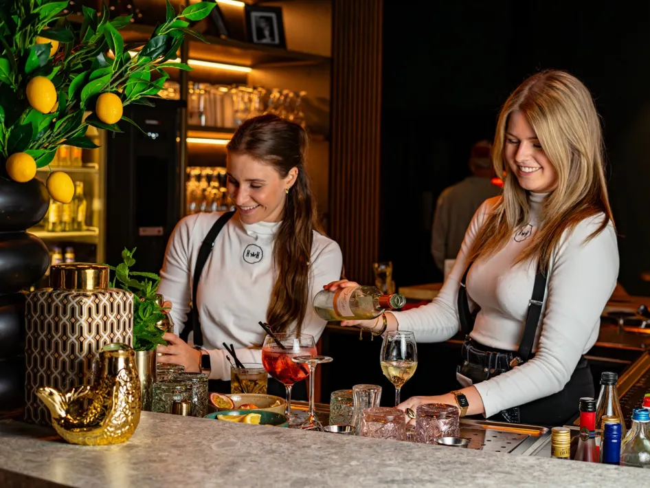 Two women behind the bar prepare cocktails with glasses, bottles and fresh garnish on the counter.